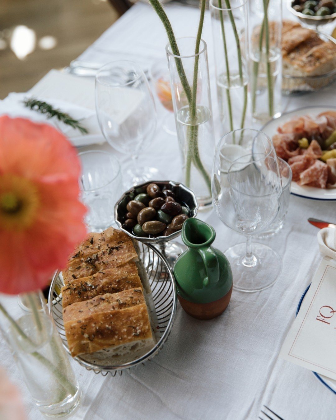 Focaccia, olives and a green jug on a white-clothed table, with a poppy in the foreground