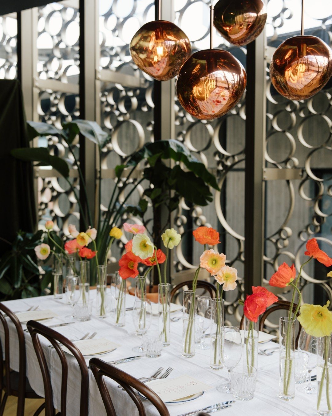 A long table set with poppies in glass vases under copper pendant lamps, in front of the patterned screens